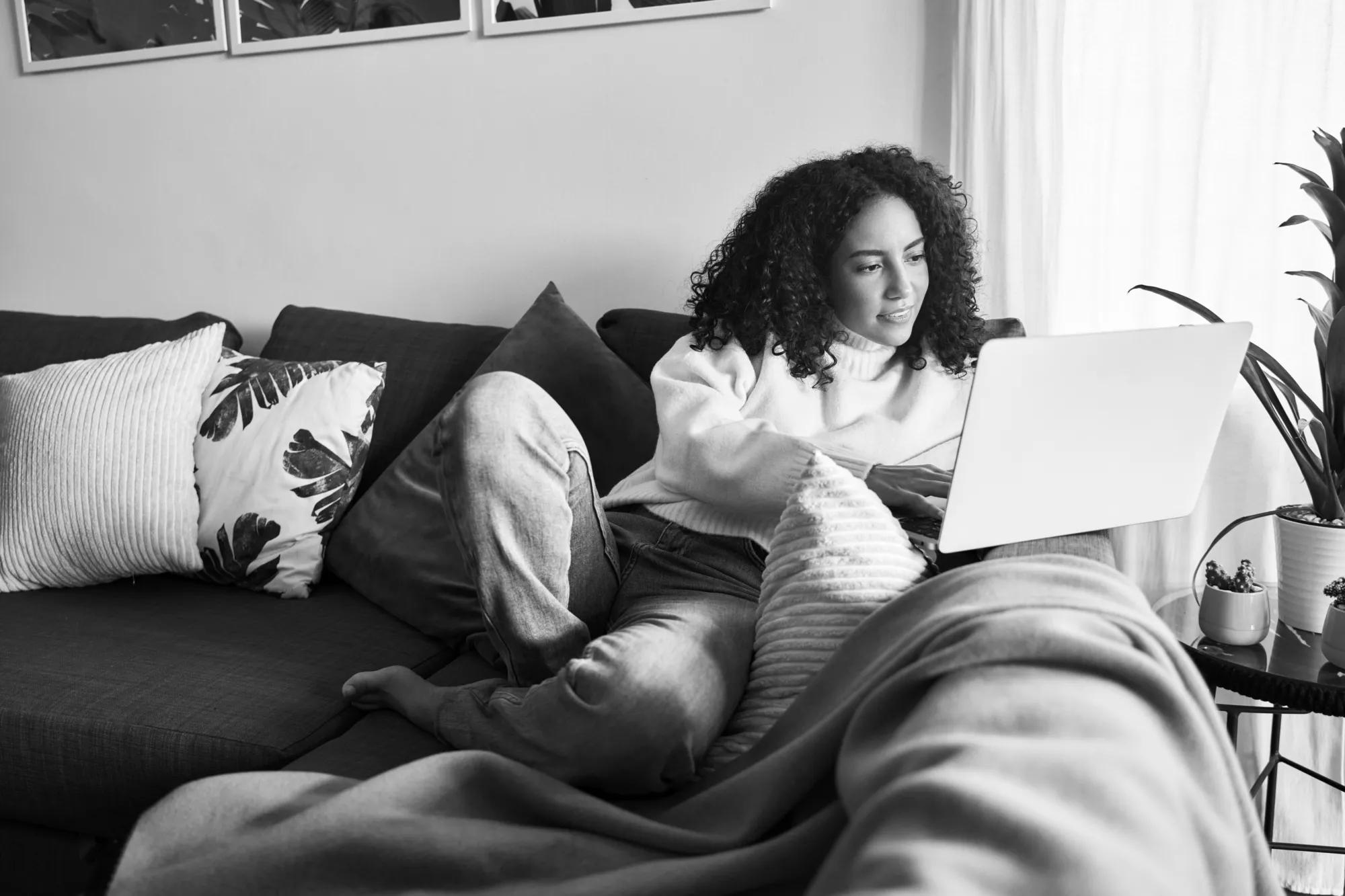 Latin young woman sitting on couch using laptop at home surfing, doing ecommerce shopping, looking at computer communicating online, searching on web, watching videos or elearning.